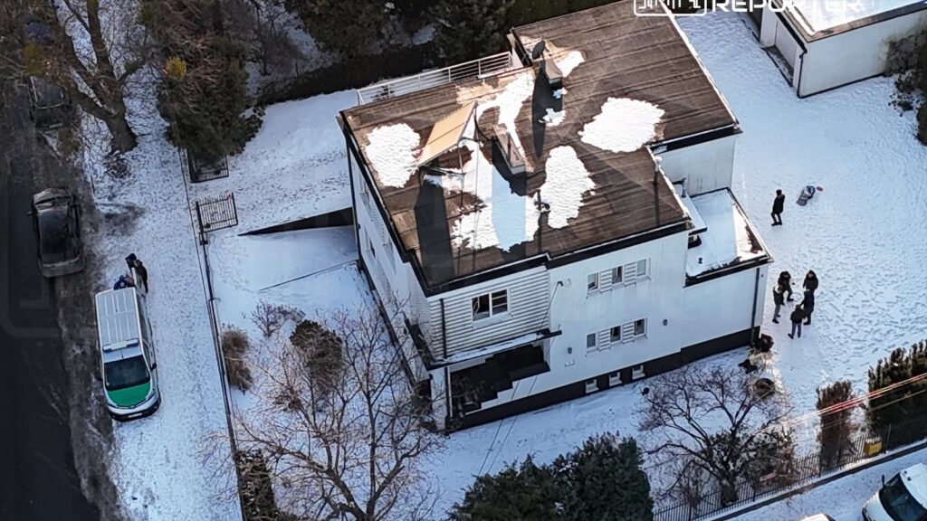 Grupa osób stoi na zaśnieżonym terenie obok białego budynku, a obok zaparkowany jest radiowóz policyjny.