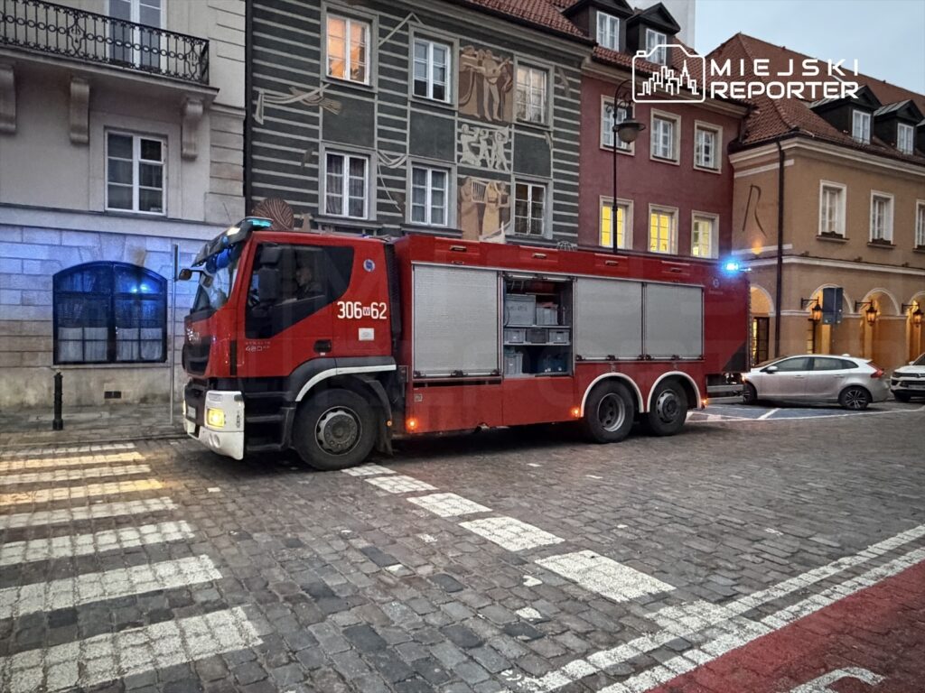 Czerwony wóz strażacki zaparkowany na brukowanej ulicy, obok budynków z kolorowymi elewacjami.