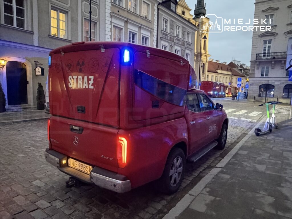 Czerwony pojazd straży z włączonymi sygnałami świetlnymi stoi na brukowanej ulicy w mieście.