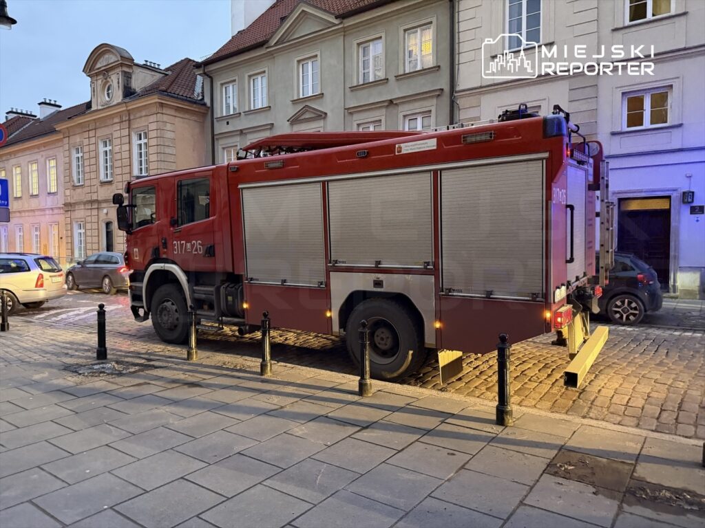 Czerwony wóz strażacki zaparkowany na wąskiej ulicy z kostki brukowej, obok zaparkowane samochody i budynki w tle.