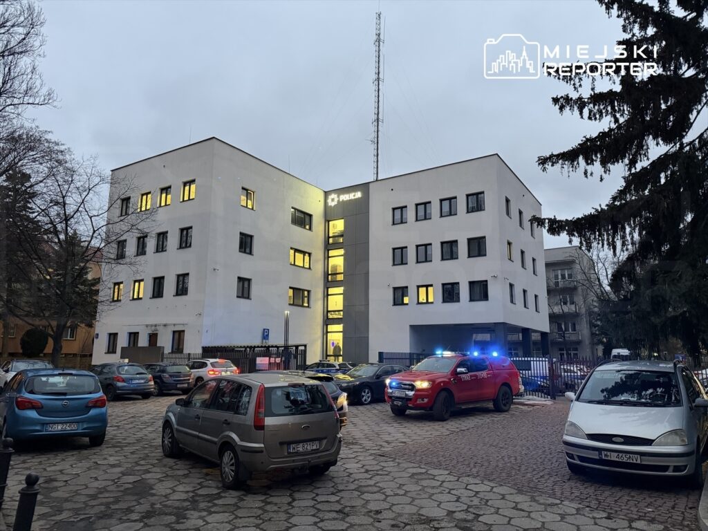 Policyjny budynek z podświetloną fasadą w otoczeniu zaparkowanych samochodów oraz wozu strażackiego z włączonymi sygnałami.