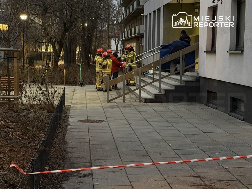 Grupa strażaków w hełmach omawia sytuację przed budynkiem mieszkalnym, w tle widoczna jest policyjna obecność.
