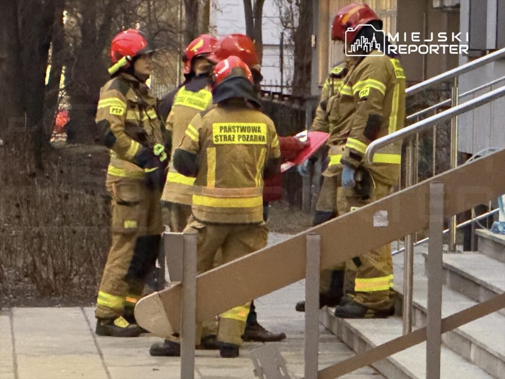 Grupa strażaków w mundurach ochronnych dyskutuje na schodach budynku, trzymając czerwony materiał ratunkowy.
