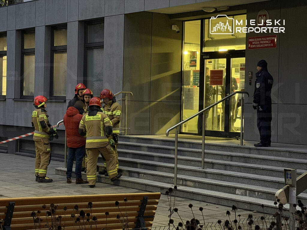 Grupa strażaków w mundurach rozmawia z osobą w czerwonej kurtce przy schodach nowoczesnego budynku.