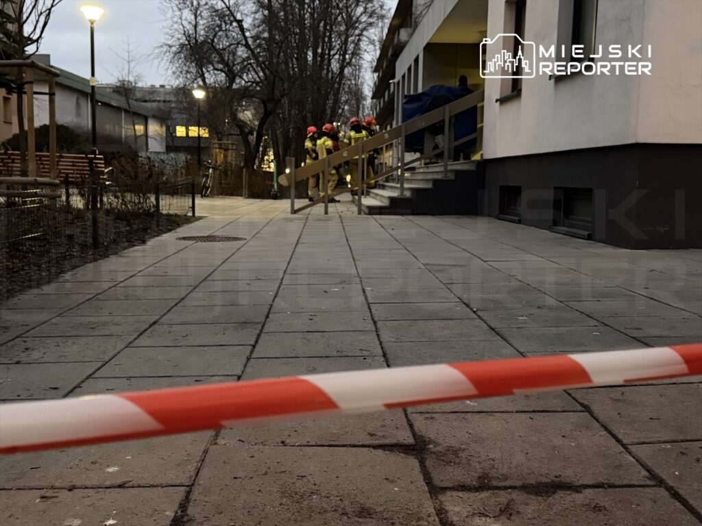 Grupa strażaków w kaskach pracuje przy schodach budynku mieszkalnego, otoczona taśmą ostrzegawczą.