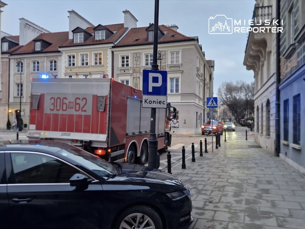 Czerwony wóz strażacki z włączonymi sygnalizatorami parkowany przy znaku drogowym „Koniec”. W tle widoczne budynki.