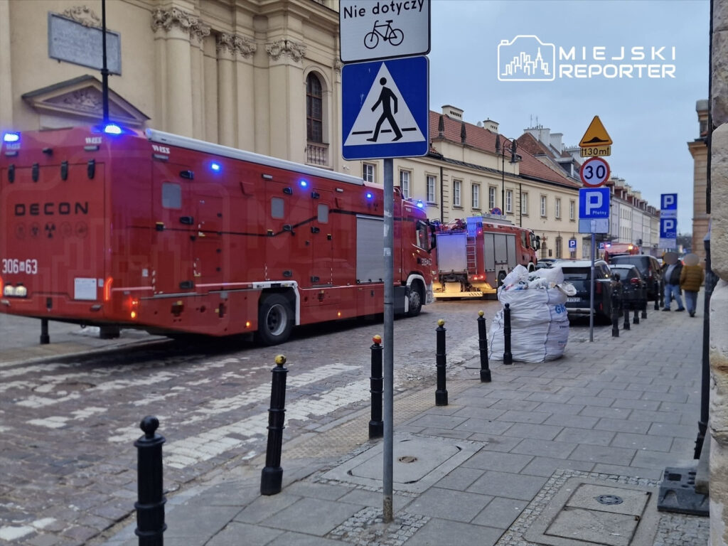 Czerwony wóz strażacki z włączonymi sygnalizatorami, zatrzymany na ulicy obok parkowanych samochodów.