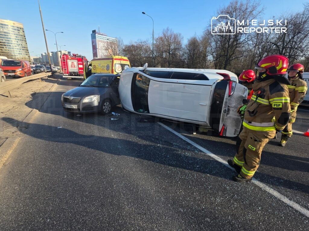 Na drodze leży przewrócony biały samochód, obok niego stoi czarny pojazd. W tle widać strażaków w mundurach, którzy oceniają sytuację.