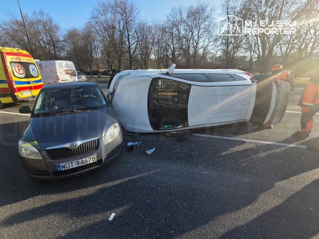 Na drodze leży przewrócony biały samochód, obok niego stoi szary pojazd, a w tle widać karetki i osoby w odblaskowych kamizelkach.