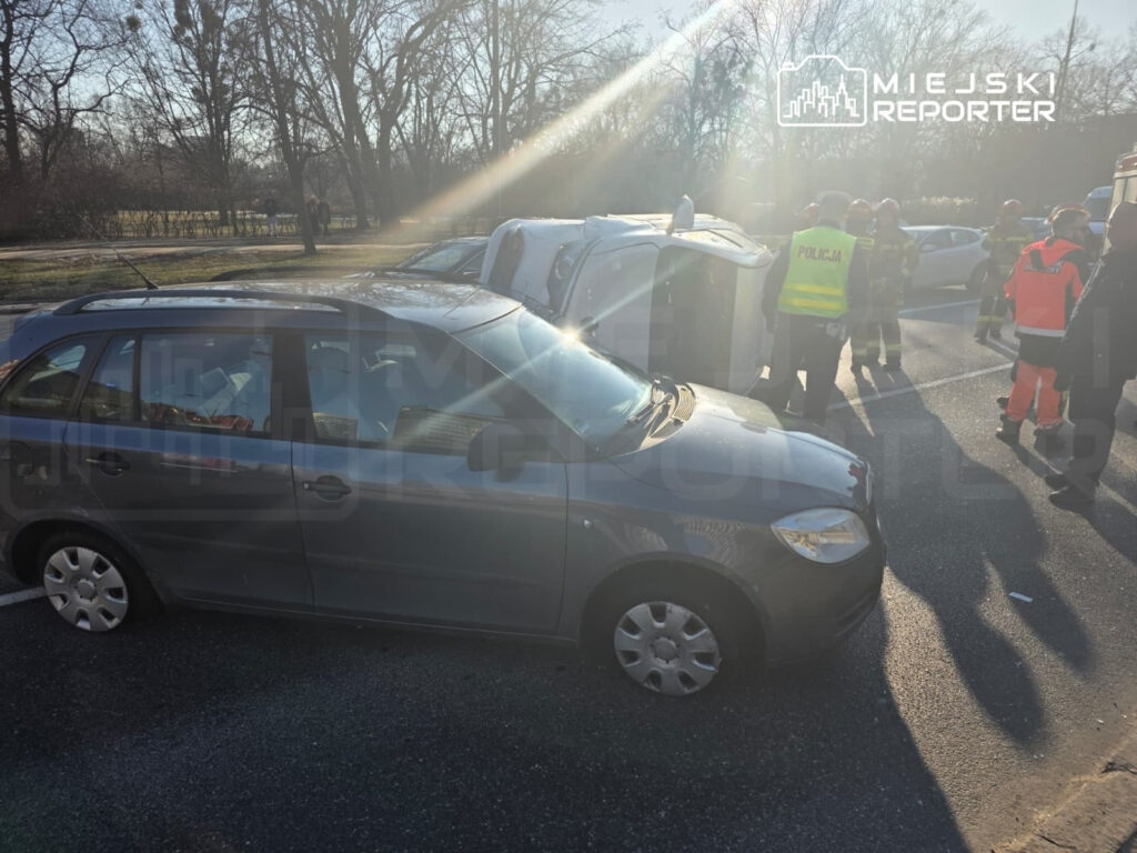 W tle widać przewrócony samochód oraz grupę ratowników i policjantów na drodze.