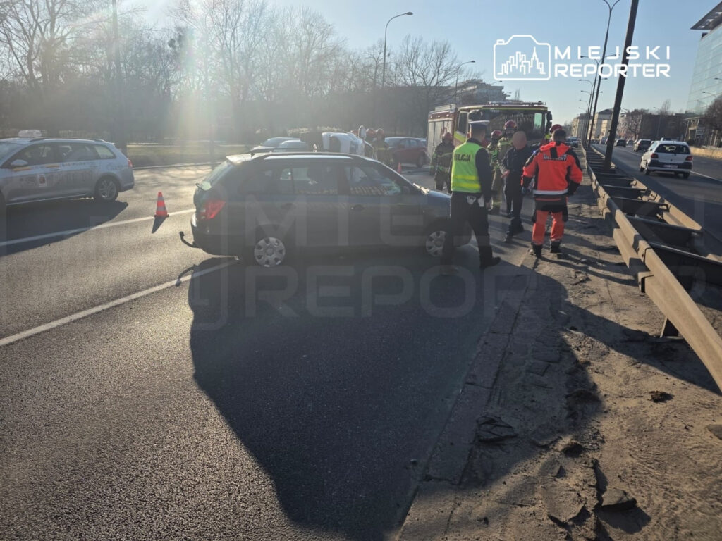 Na drodze stoi czarny samochód z uszkodzeniem, wokół niego grupka ratowników i policjantów.