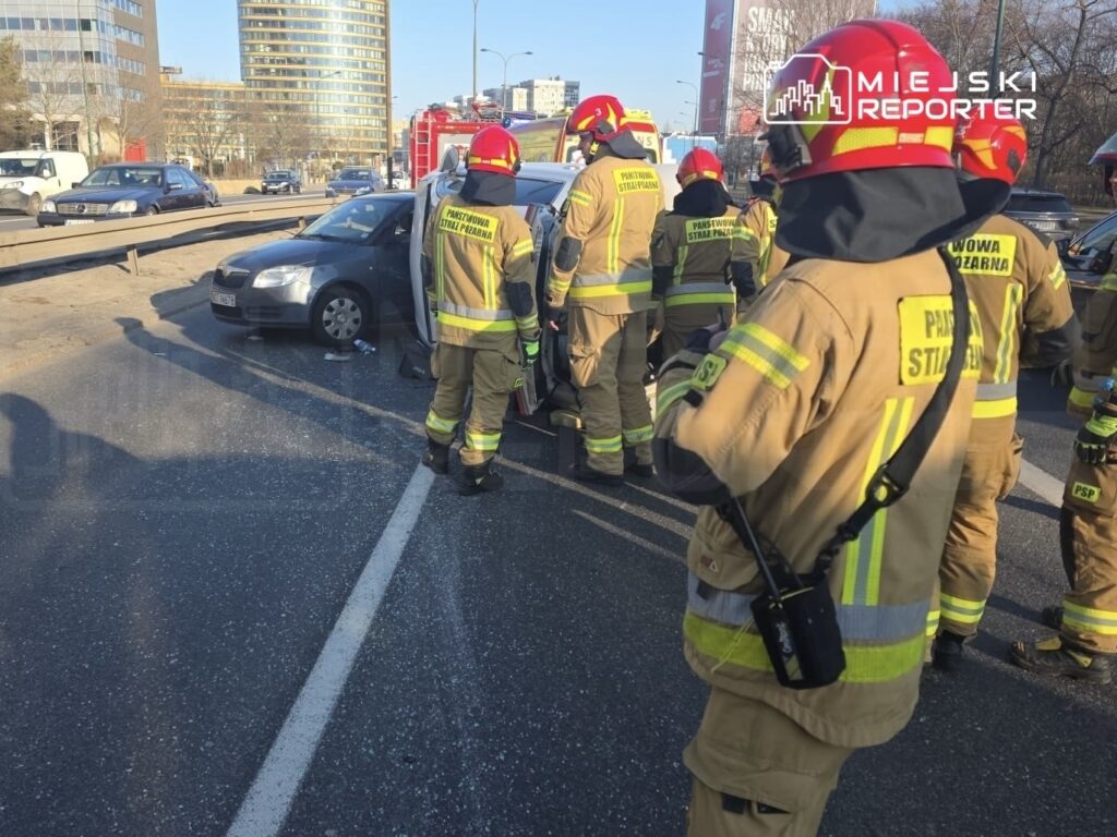 Grupa strażaków w mundurach z logo PSP udziela pomocy po wypadku drogowym na miejskiej ulicy.