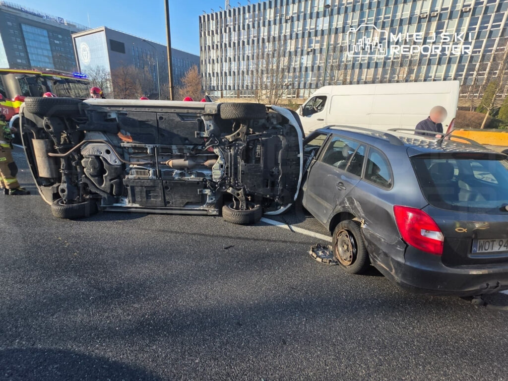 Na drodze leży przewrócony samochód osobowy, obok niego stoi uszkodzony drugi pojazd, a w tle widać nowoczesne budynki.