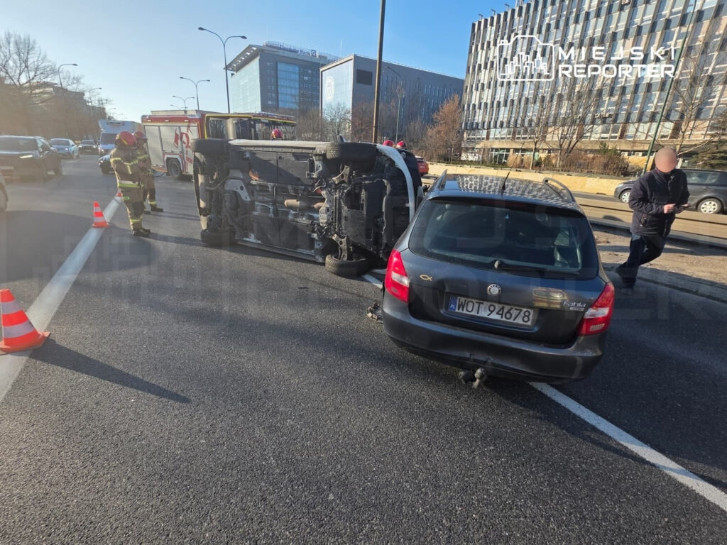 Na drodze leży przewrócony samochód, obok niego stoją strażacy w uniformach, a w tle widać ruch uliczny.