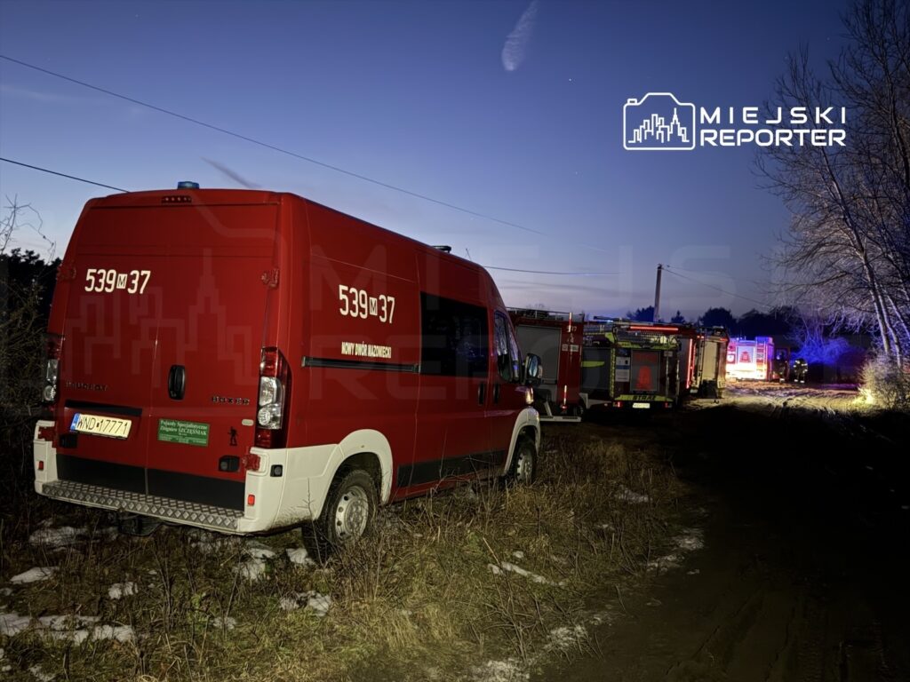 Czerwony samochód strażacki zaparkowany na poboczu drogi w lesie, w tle widoczne inne pojazdy ratunkowe i migające światła.