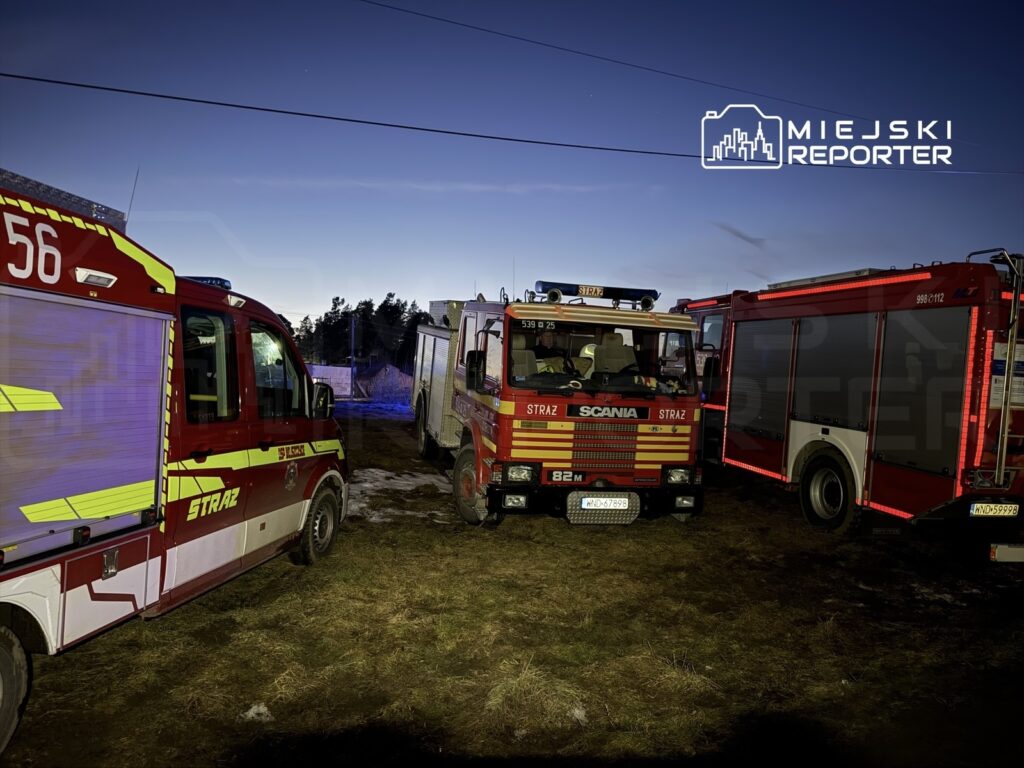 Trzy samochody strażackie zaparkowane na trawie w ciemnym otoczeniu z widocznym niebem.