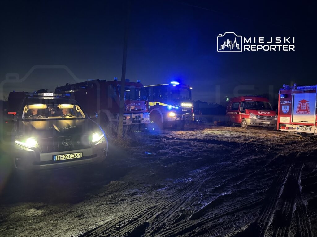 Policyjny samochód z włączonymi światłami, obok pojazdy straży pożarnej, na ciemnej drodze w nocy.
