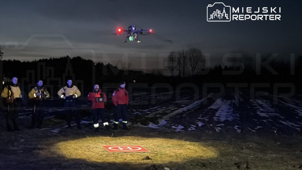 Grupa ratowników w odzieży ochronnej obserwuje dron nad oświetlonym polem z logo lądowiska.