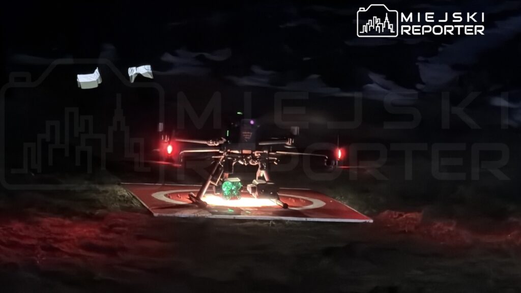 Dron z zielonym oświetleniem ląduje na oznakowanym miejscu w ciemnym otoczeniu.