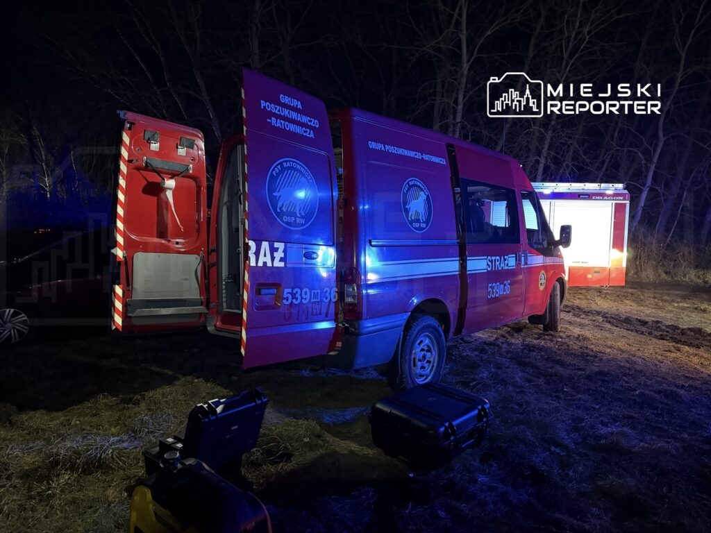 Czerwony samochód ratunkowy z otwartymi drzwiami stoi na leśnej polanie w nocy, otoczony przez drzewa.