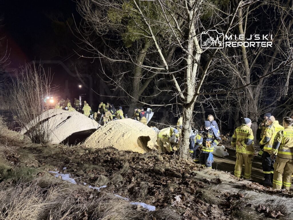 Grupa strażaków pracuje nocą przy hałdach piasku w pobliżu drzew, organizując akcję ratunkową.