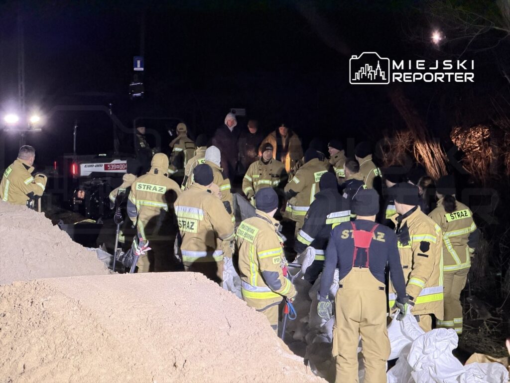 Grupa strażaków w mundurach roboczych pracuje nocą nad zabezpieczeniem terenu, transportując worki z piaskiem.