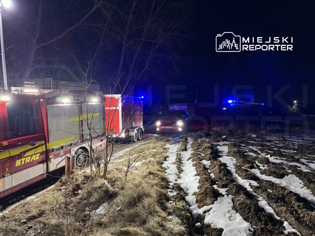 W nocy, w terenie z zaśnieżoną ziemią, stoją dwa pojazdy straży pożarnej z włączonymi sygnałami świetlnymi.