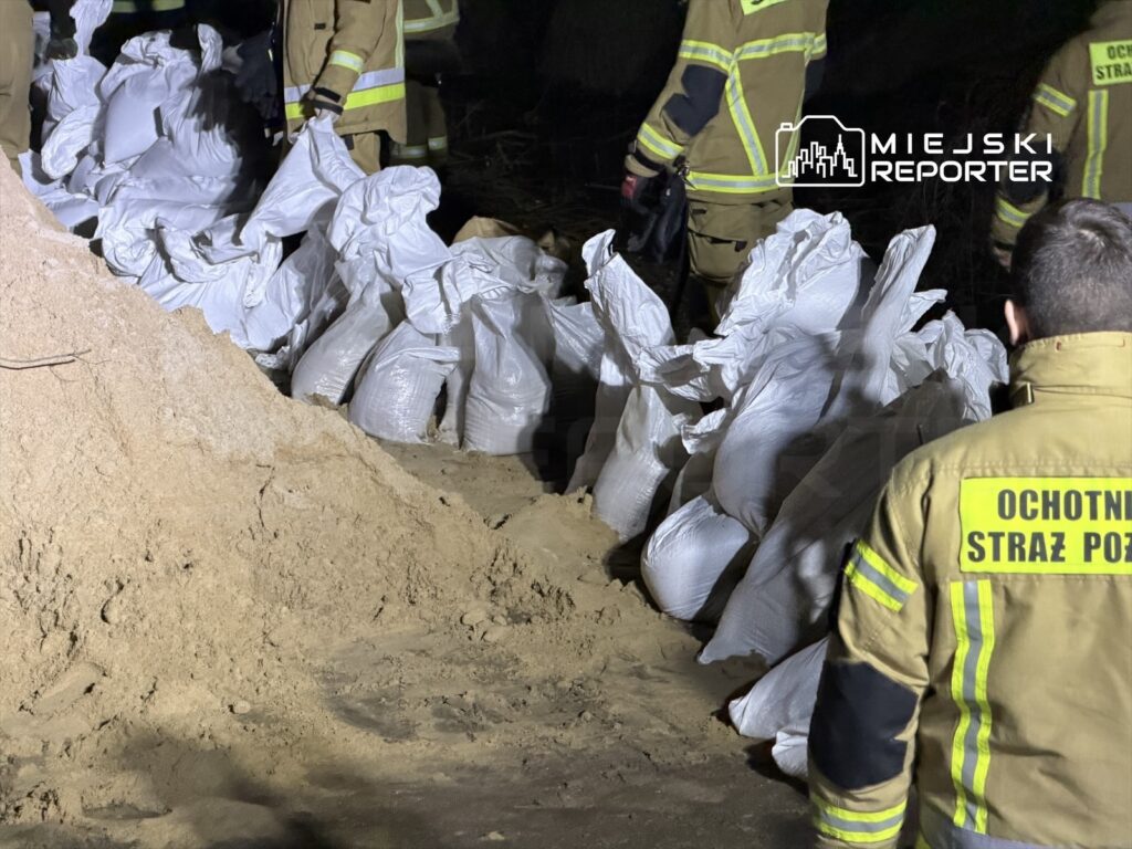 Strażacy w mundurach przygotowują worki z piaskiem w ciemnym terenie, otoczeni przez zasypane piaskiem miejsce.