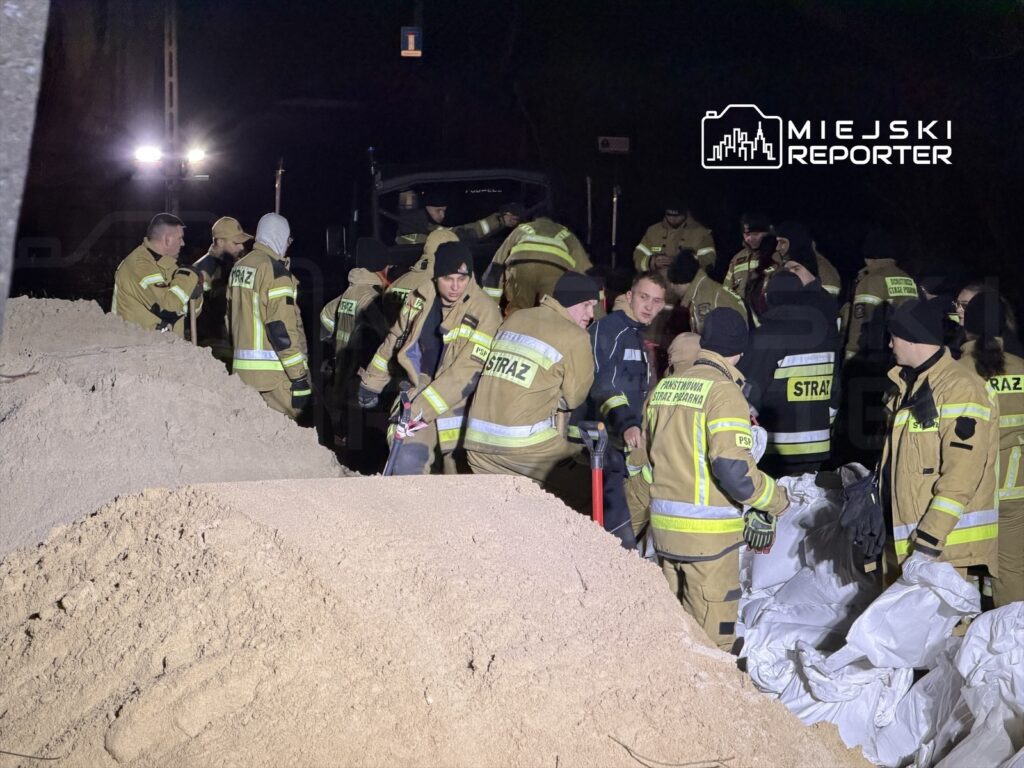 Grupa strażaków w mundurach pracuje nocą przy usuwaniu piasku i organizacji sprzętu ratunkowego.