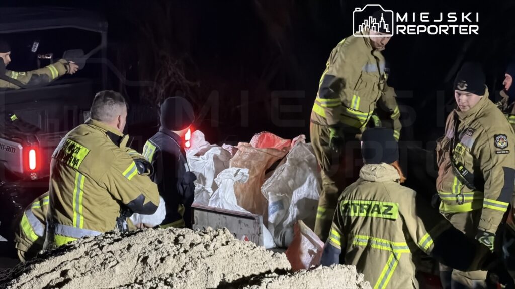 Grupa strażaków w mundurach przeszukuje teren w nocy, otaczając worki z piaskiem.