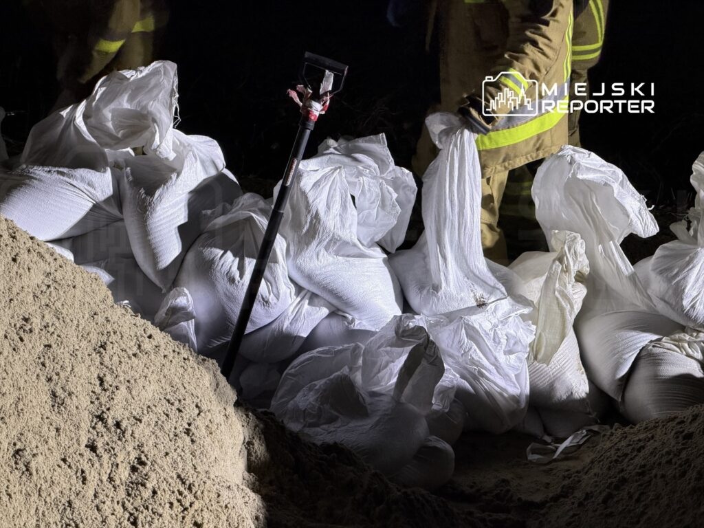 Grupa strażaków w mundurach pracuje nocą, układając białe worki z piaskiem w wykopie.