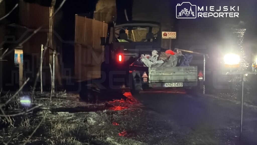 Ciężarówka z przyczepą załadowaną workami stoi na mokrej nawierzchni nocą, oświetlona reflektorami.