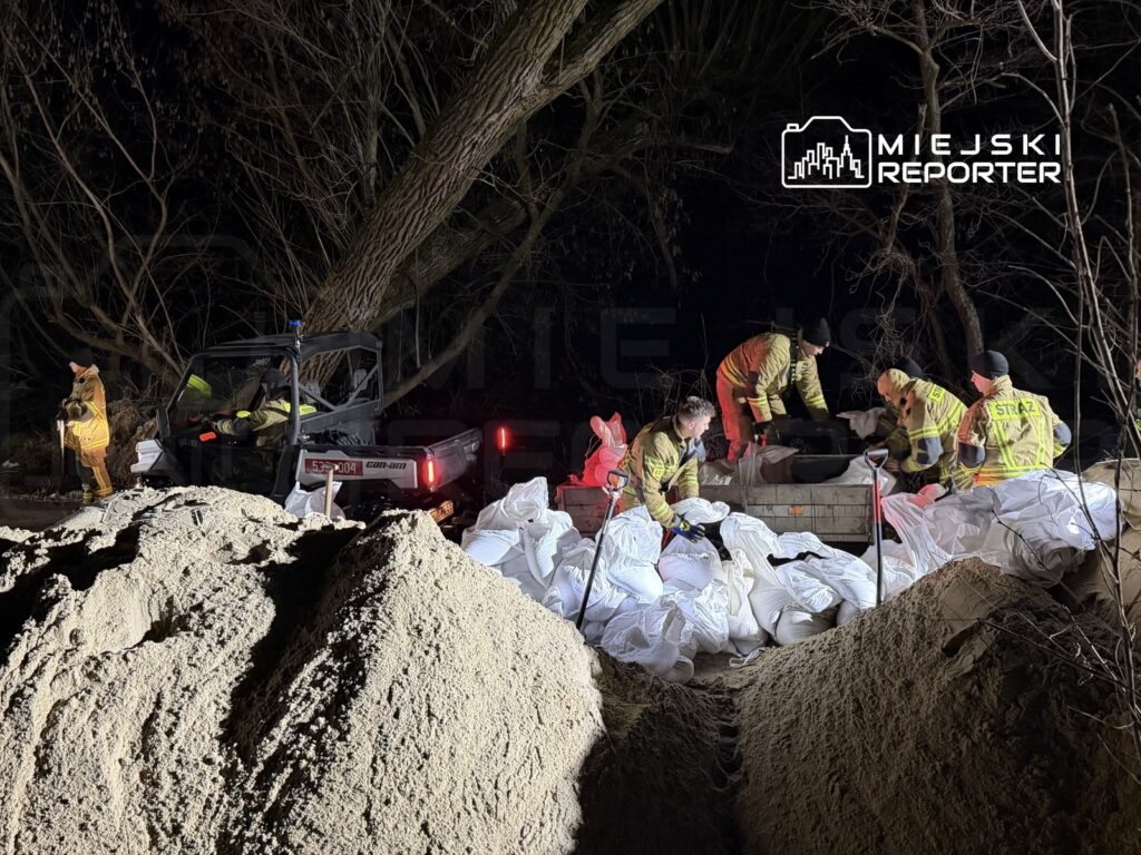 Grupa strażaków pracuje nocą, zabezpieczając teren piaskiem i workami, obok pojazdu terenowego w lesie.