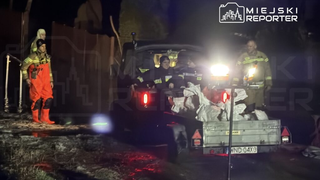 Strażacy w pomarańczowych kombinezonach pracują przy załadunku worków na przyczepę w ciemnej okolicy po powodzi.