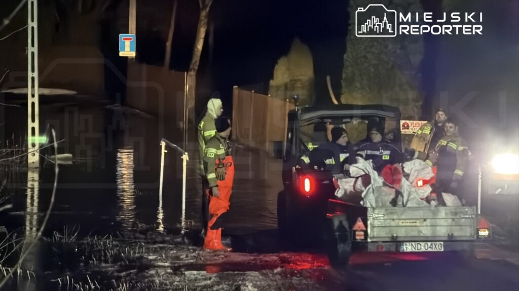 Grupa ratowników w odzieży ochronnej pracuje w zalanym terenie, przy pojeździe z workami. W tle widoczne znaki drogowe.