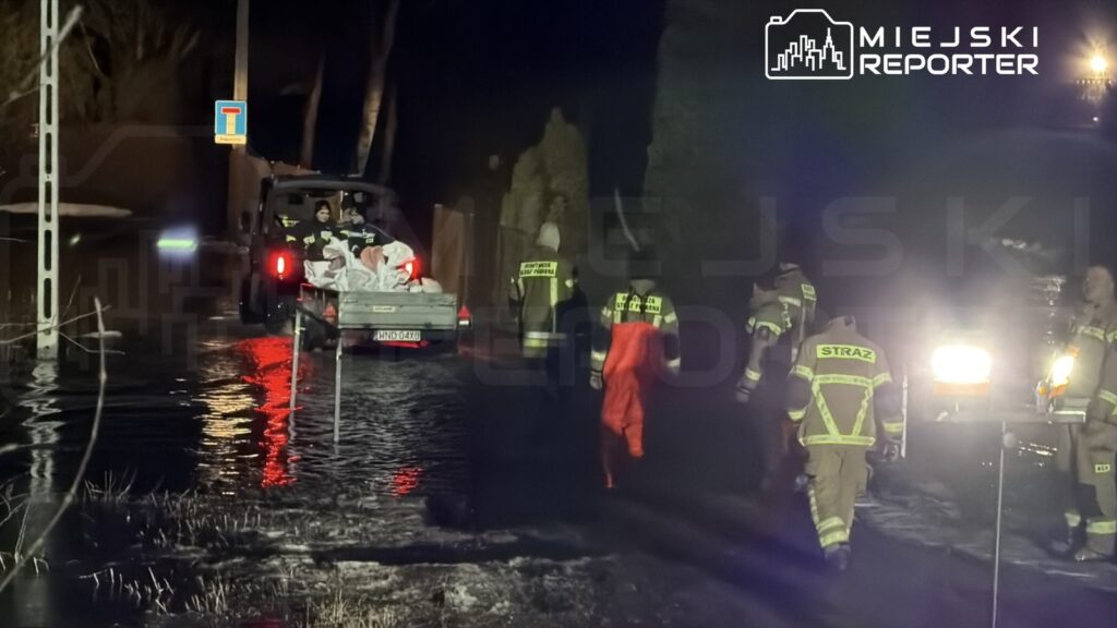 Strażacy w odzieży ochronnej przeprowadzają akcję ratunkową w terenie zalanym wodą, przy użyciu pojazdu terenowego.