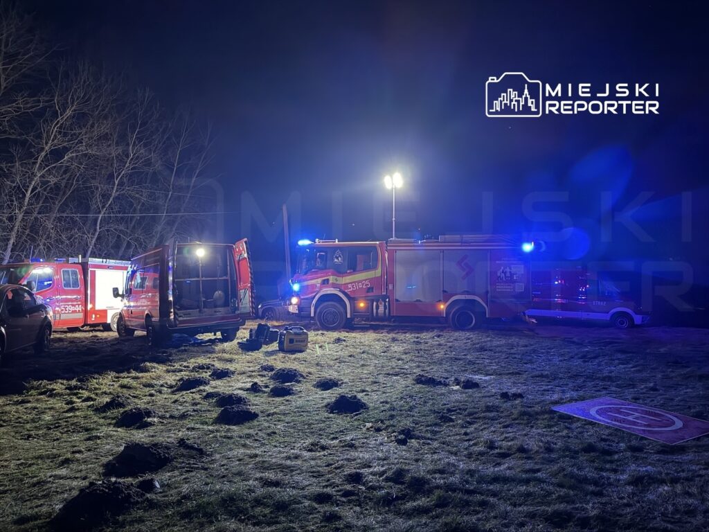 Zespół ratunkowy pracuje w nocy przy wozach strażackich na oświetlonym terenie z widocznymi wykopami w ziemi.