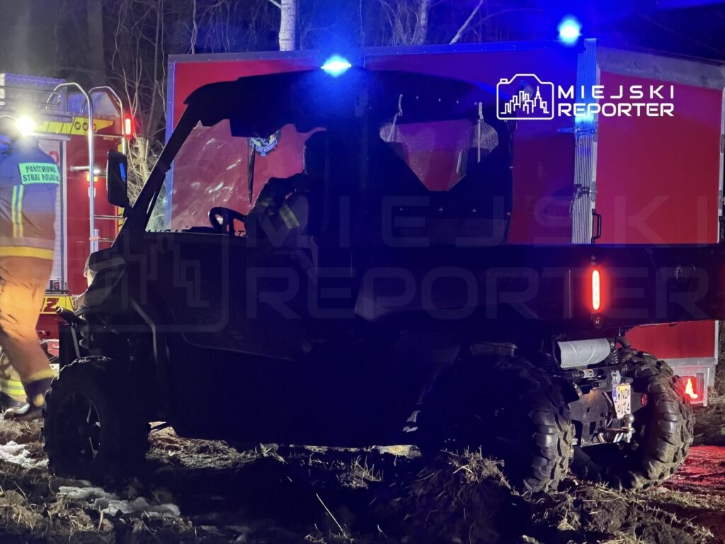 Strażak w odblaskowym mundurze przy pojazdu ratunkowym w lesie, obok terenowego pojazdu z włączonymi światłami.