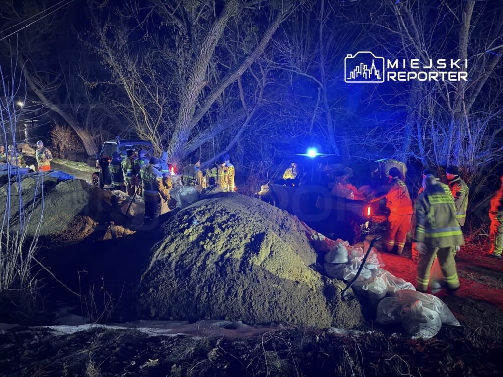 Grupa ratowników w odblaskowych strojach transportuje worki z piaskiem w ciemnym, leśnym otoczeniu, oświetlonym przez niebieskie i czerwone światła pojazdów.