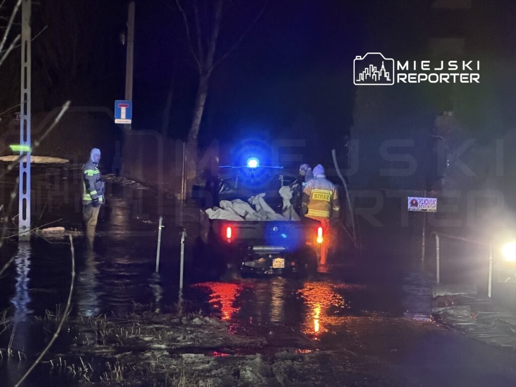 Strażacy w odblaskowych mundurach w nocy pracują przy zalanej drodze, zabezpieczając teren workami z piaskiem.
