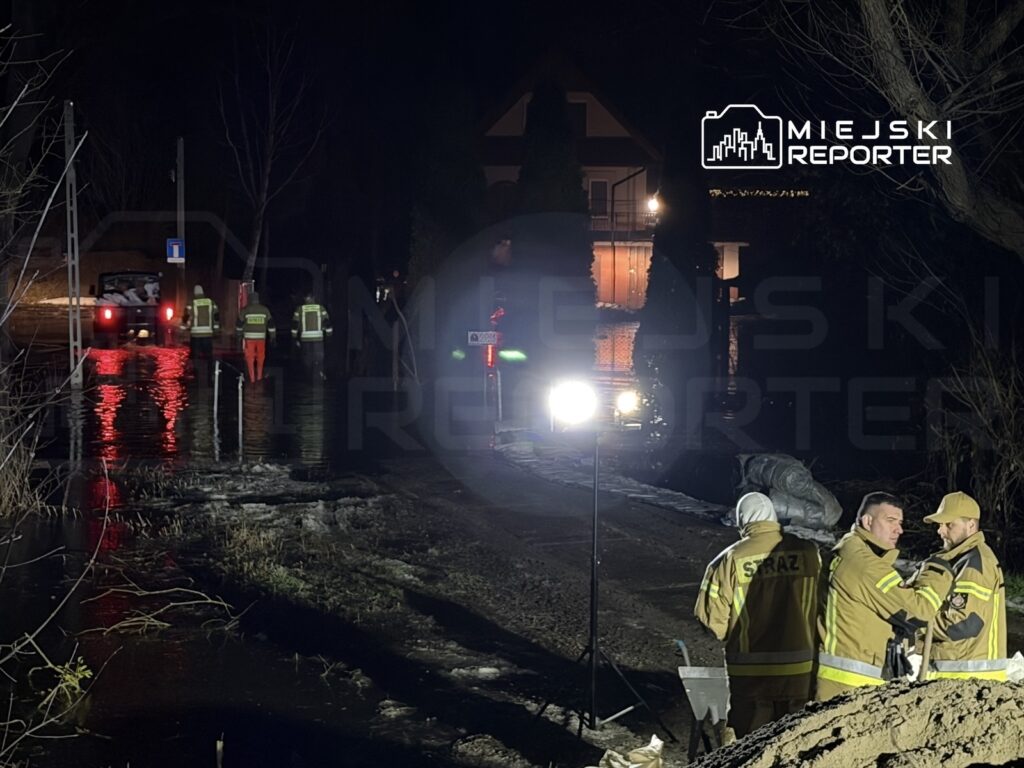 Grupa strażaków w odblaskowych kamizelkach pracuje w nocy w zalanym terenie, obok samochodów i sprzętu ratunkowego.