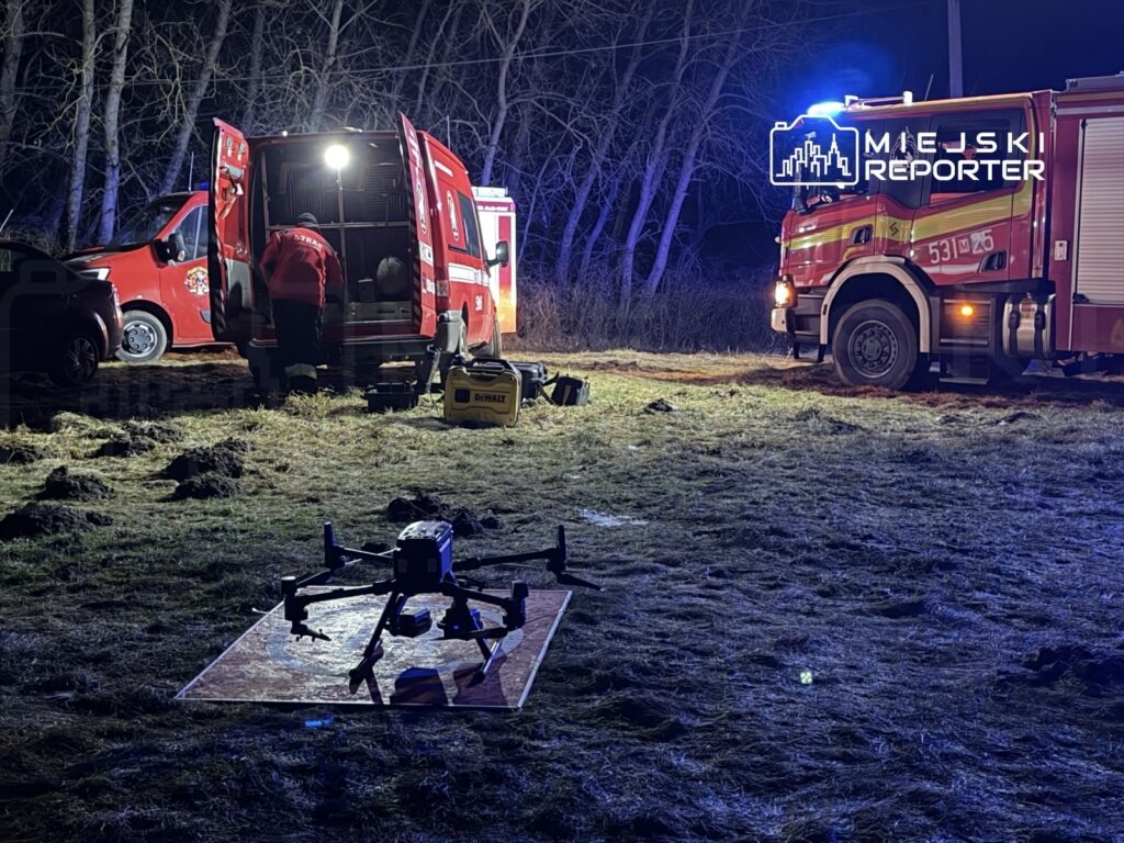 Strażacy przy samochodzie ratunkowym przygotowują drona do akcji w terenie leśnym w nocy.