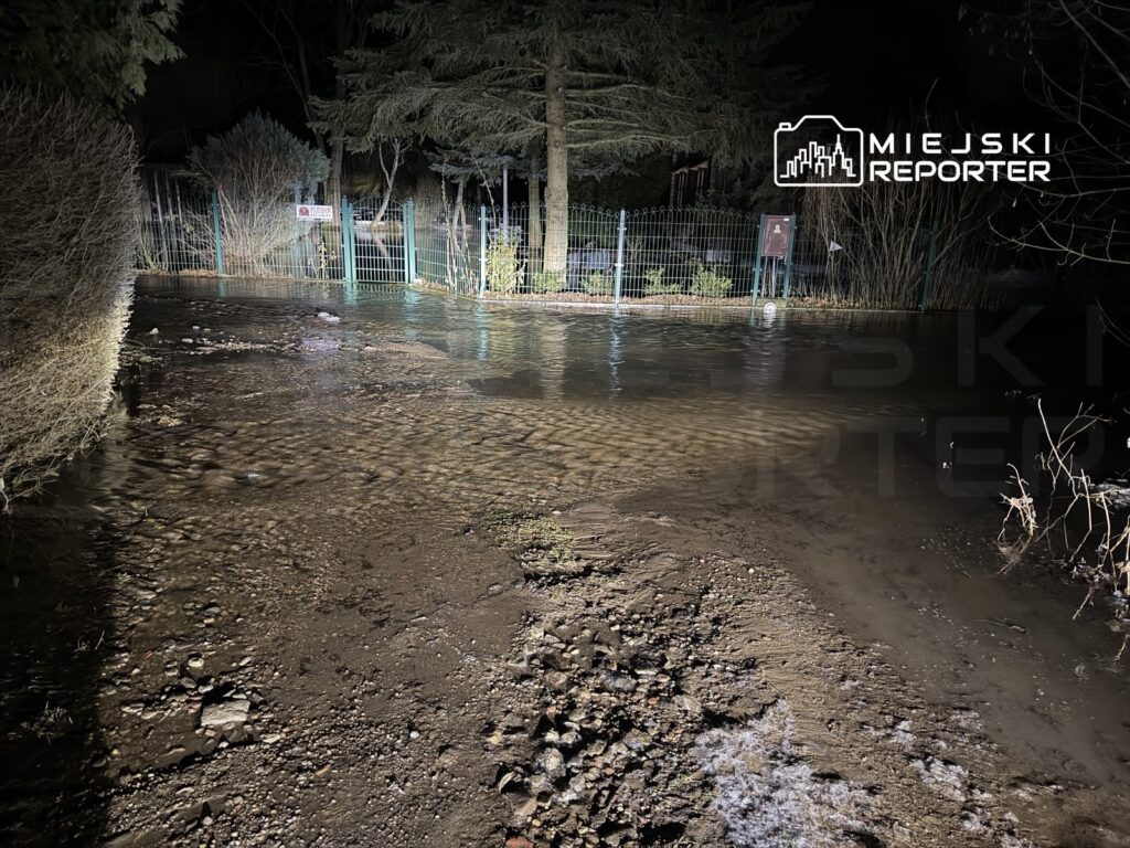 Zalany teren z wodą sięgającą do kostek, otoczony ogrodzeniem i drzewami, w nocy przy oświetleniu.