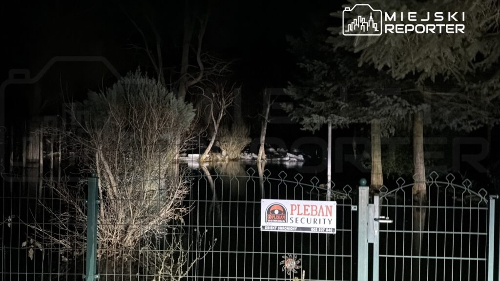 Zielony płot z bramą zabezpieczoną znakiem "PLEBAN SECURITY" w otoczeniu ciemnego lasu i zarysu wody nocą.