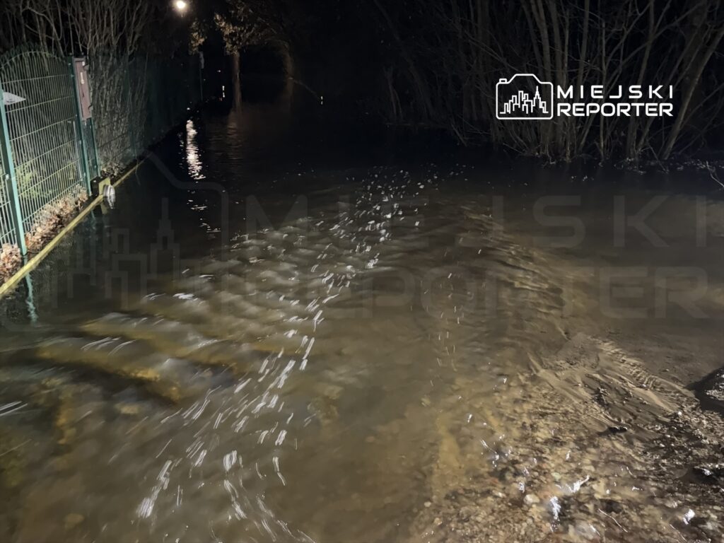 Zalany chodnik w ciemnym otoczeniu, woda sięga do krawędzi ogrodzenia.