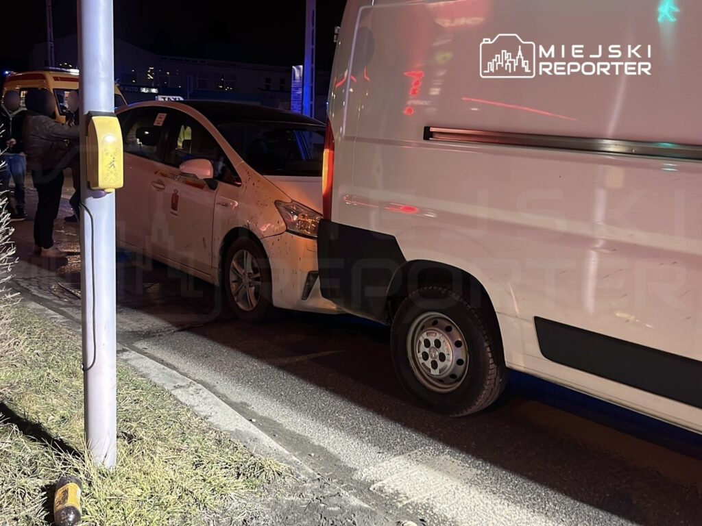 Kierowca taksówki rozmawia z pasażerem obok zaparkowanego furgonu na oświetlonej ulicy nocą.