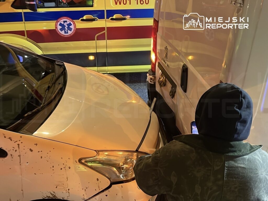 Mężczyzna w kapturze stoi obok zderzonego samochodu, a w tle widoczna karetka pogotowia.