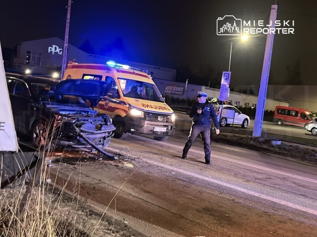 Policyjny funkcjonariusz bada wypadek drogowy przy uszkodzonym samochodzie, obok stoi karetka pogotowia.