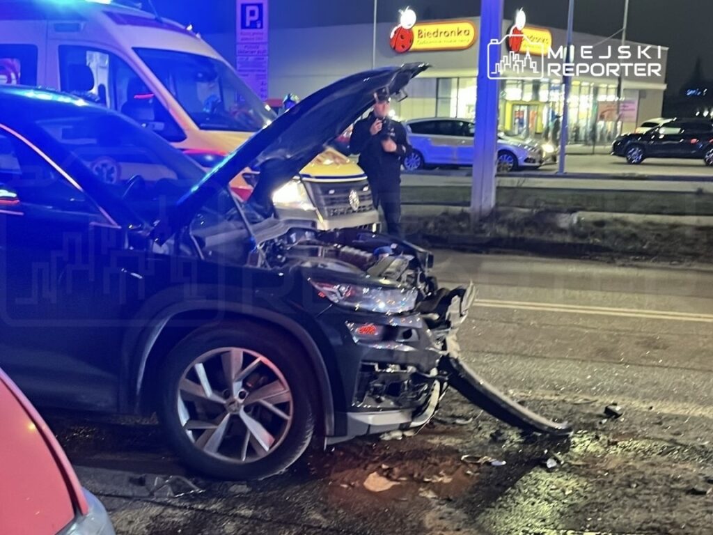 Uszkodzony samochód osobowy z otwartą maską stoi na drodze, obok interweniuje policjant, w tle widoczny sklep.