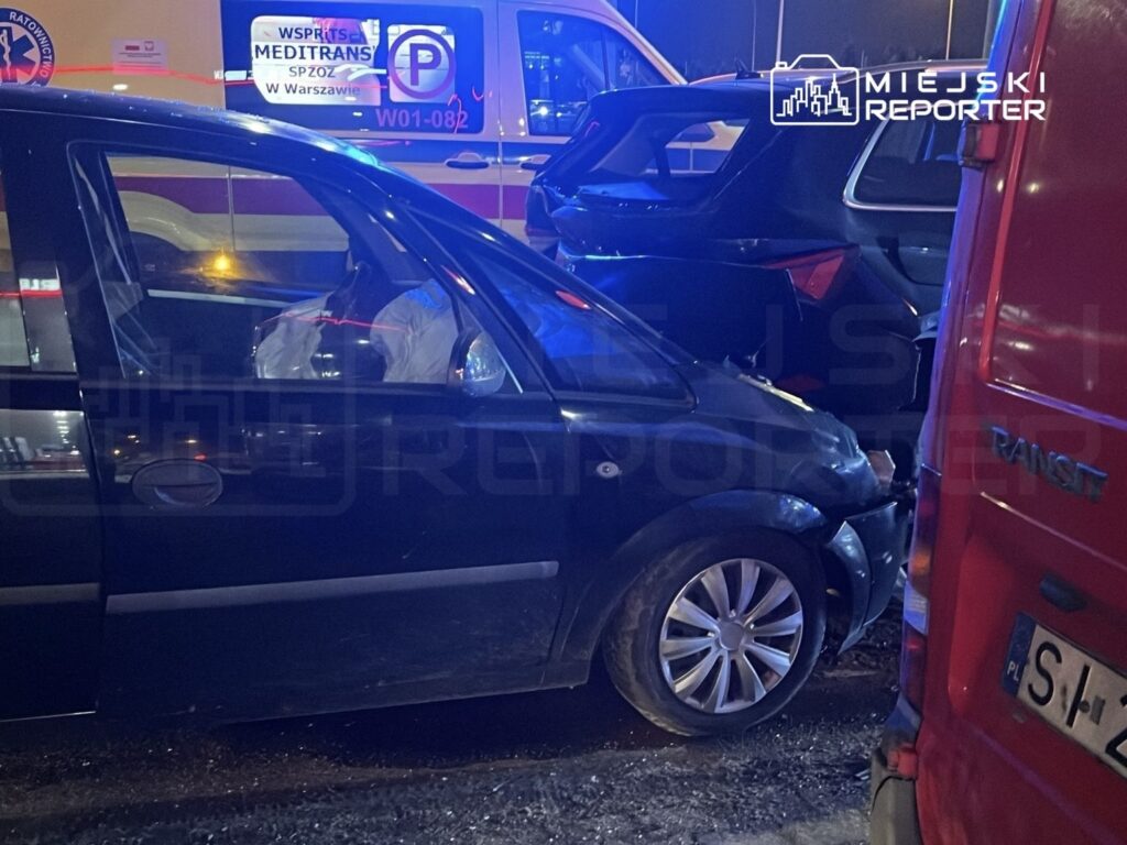Czarny samochód osobowy z uszkodzoną karoserią stoi obok innych pojazdów w nocy, w tle widać karetki pogotowia.
